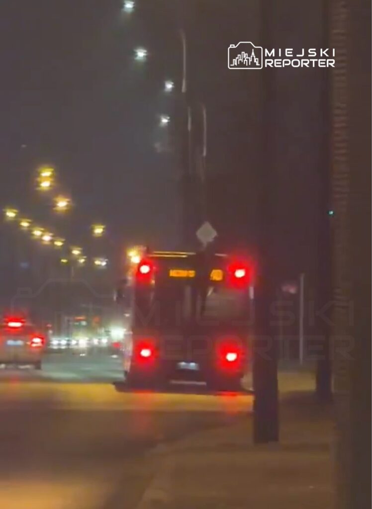 Autobus z włączonymi światłami tylnymi porusza się po oświetlonej ulicy nocą, mijając inne pojazdy.