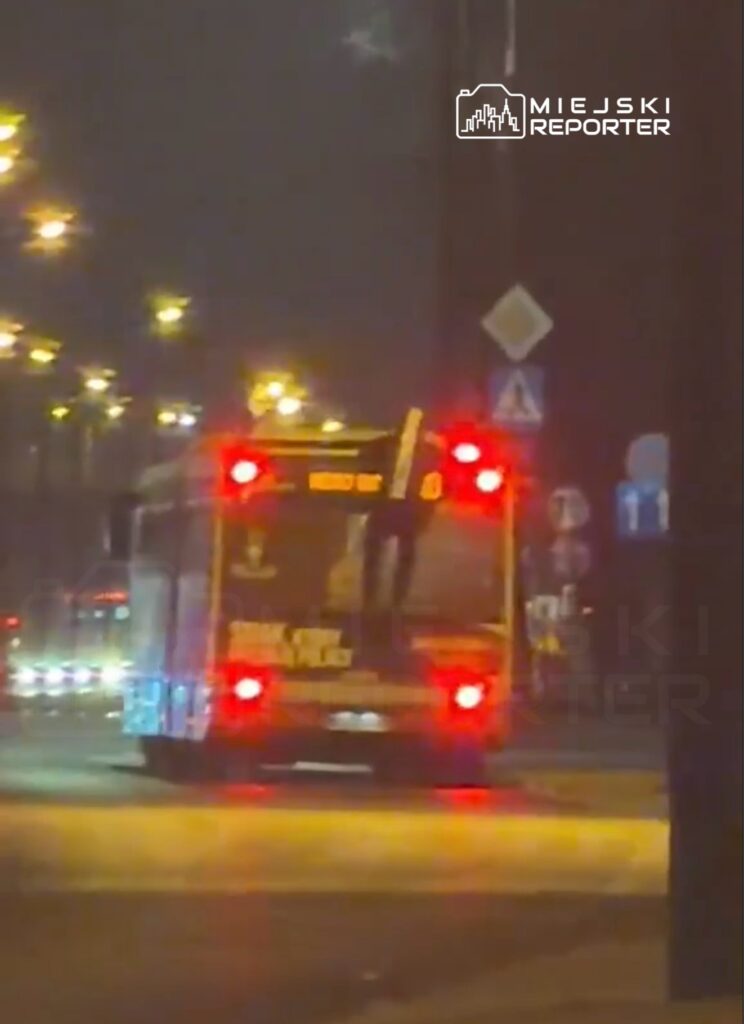 Czerwony autobus z włączonymi światłami hamowania skręca w ulicę nocą, w tle widoczne inne pojazdy i latarnie.