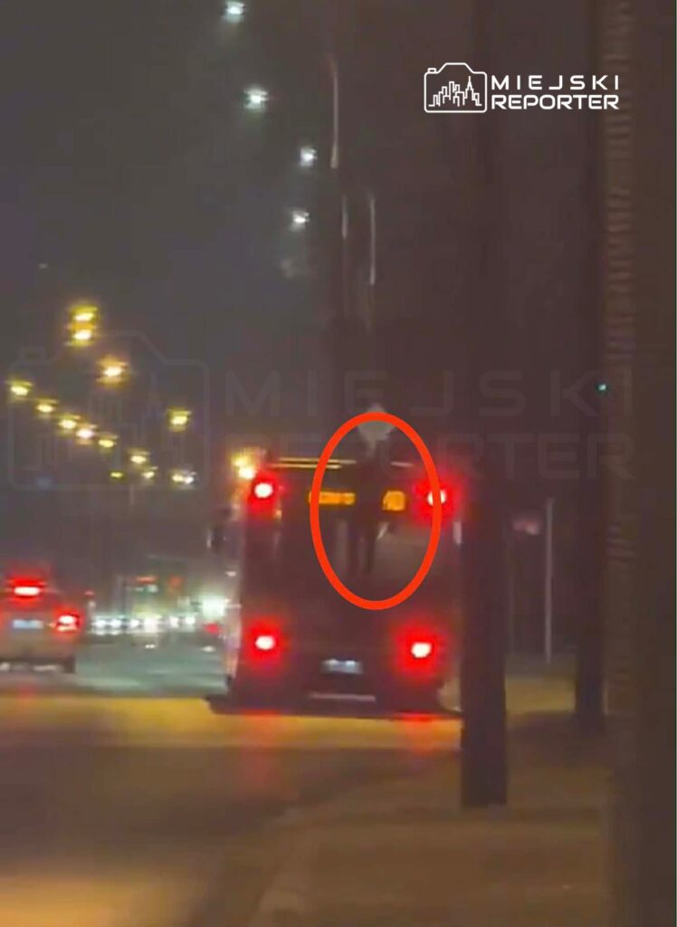 Osoba stoi na tylnym dachu autobusu miejskiego, podczas gdy pojazd porusza się po oświetlonej ulicy nocą.