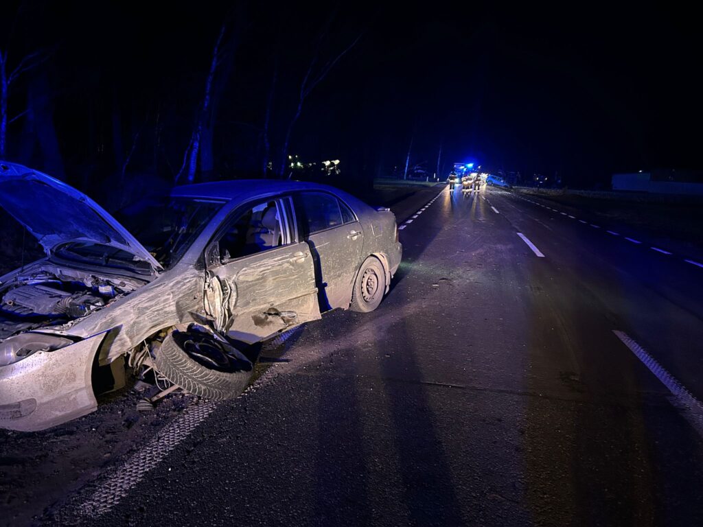 Uszkodzony samochód osobowy z uniesioną maską stoi na poboczu drogi, w tle widoczne niebieskie światła radiowozów.