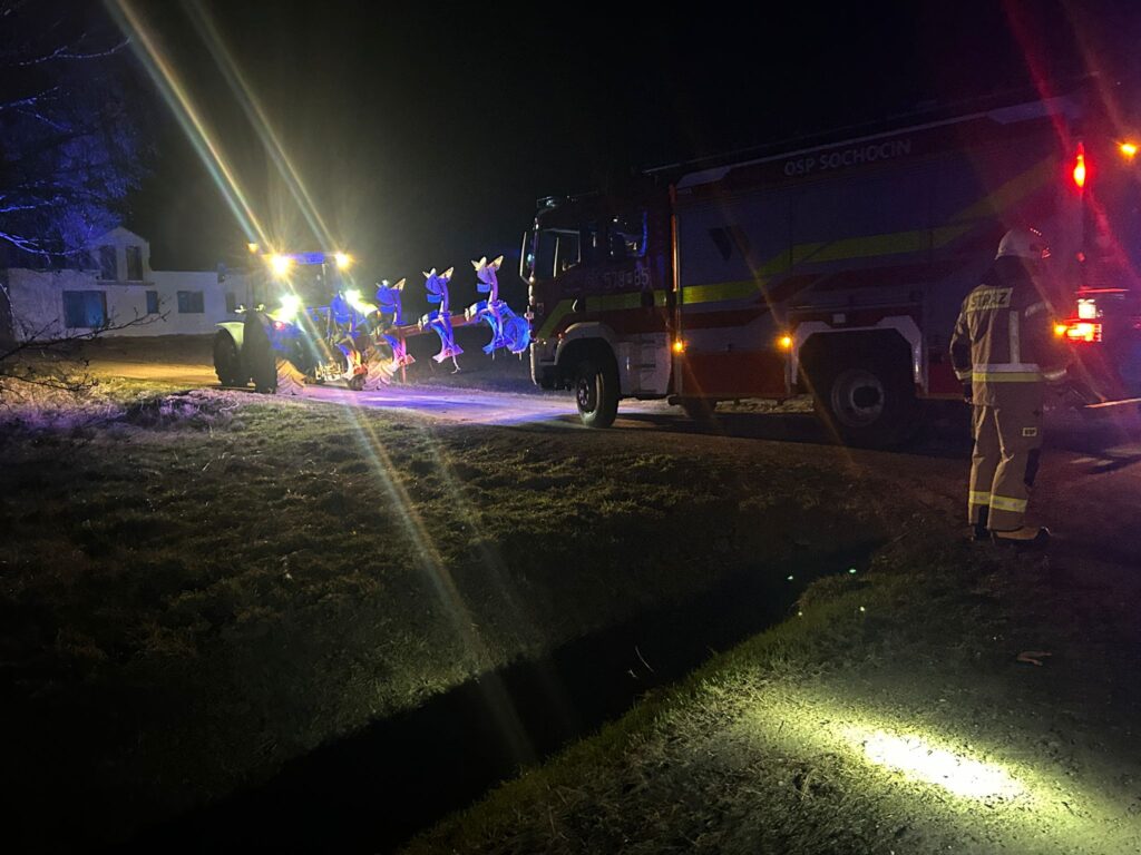 Traktor z oświetleniem roboczym odśnieża drogę w nocy, obok stoi wóz strażacki i strażak w mundurze.