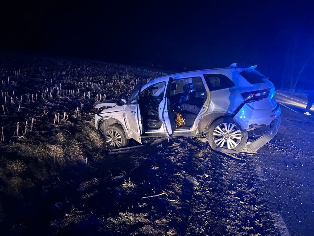 Uszkodzony samochód osobowy stoi na poboczu drogi, z otwartymi drzwiami, w ciemnej okolicy oświetlonej niebieskimi światłami.