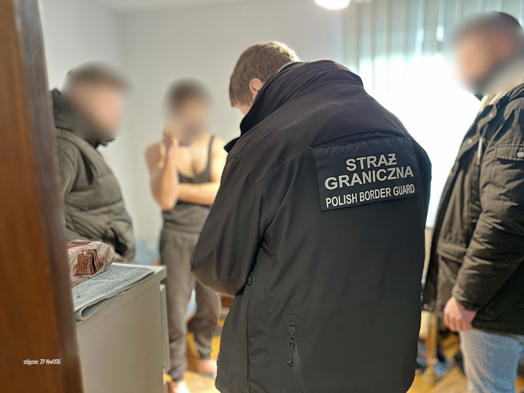 Funkcjonariusz Straży Granicznej rozmawia z dwoma mężczyznami w pomieszczeniu mieszkalnym.