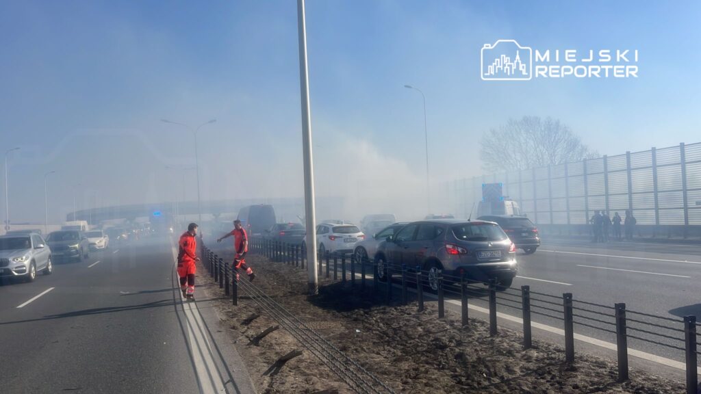 Dwaj ratownicy w czerwonych strojach sprawdzają sytuację na drodze, gdzie unosi się dym, obok stojących pojazdów.