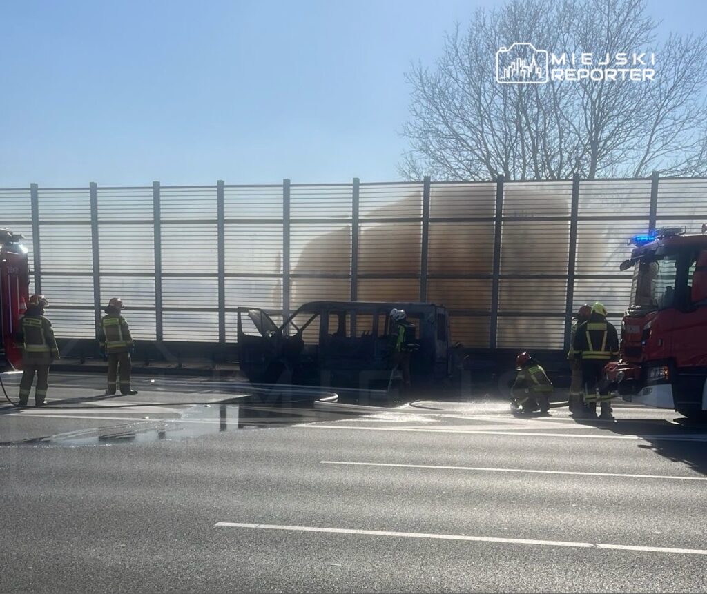 Strażacy gaszą płonący samochód na poboczu drogi, obok widoczny wóz strażacki i ogrodzenie akustyczne.