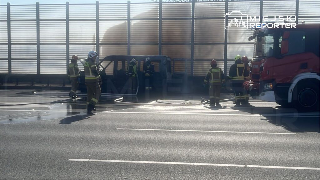 Grupa strażaków gasi pożar uszkodzonego pojazdu na drodze, obok stoi wóz strażacki. W tle widoczna bariera dźwiękochłonna.