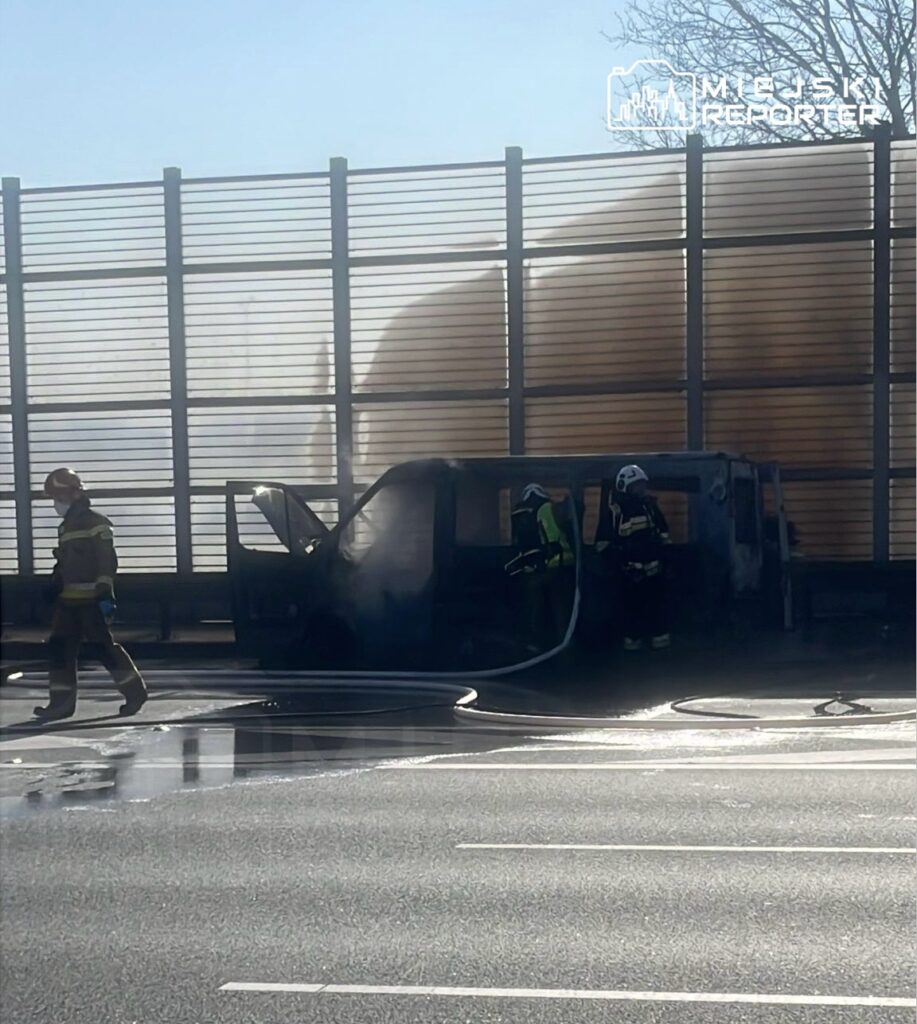 Strażacy gaszą palący się pojazd na poboczu drogi, w tle widać ogrodzenie dźwiękochłonne.