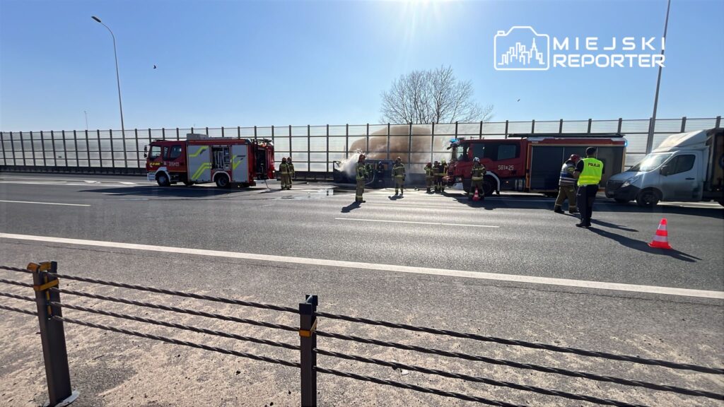 Strażacy gaszą pożar samochodu na autostradzie, obok stoją wozem strażackim i policjant w kamizelce odblaskowej.