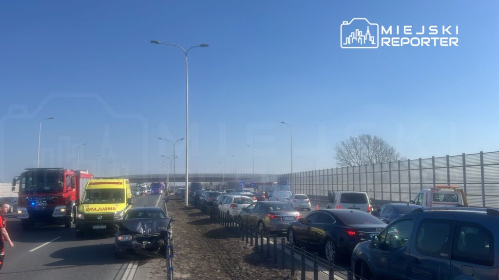 Na drodze widać zator spowodowany wypadkiem, w którym uczestniczą pojazdy ratunkowe i uszkodzony samochód.