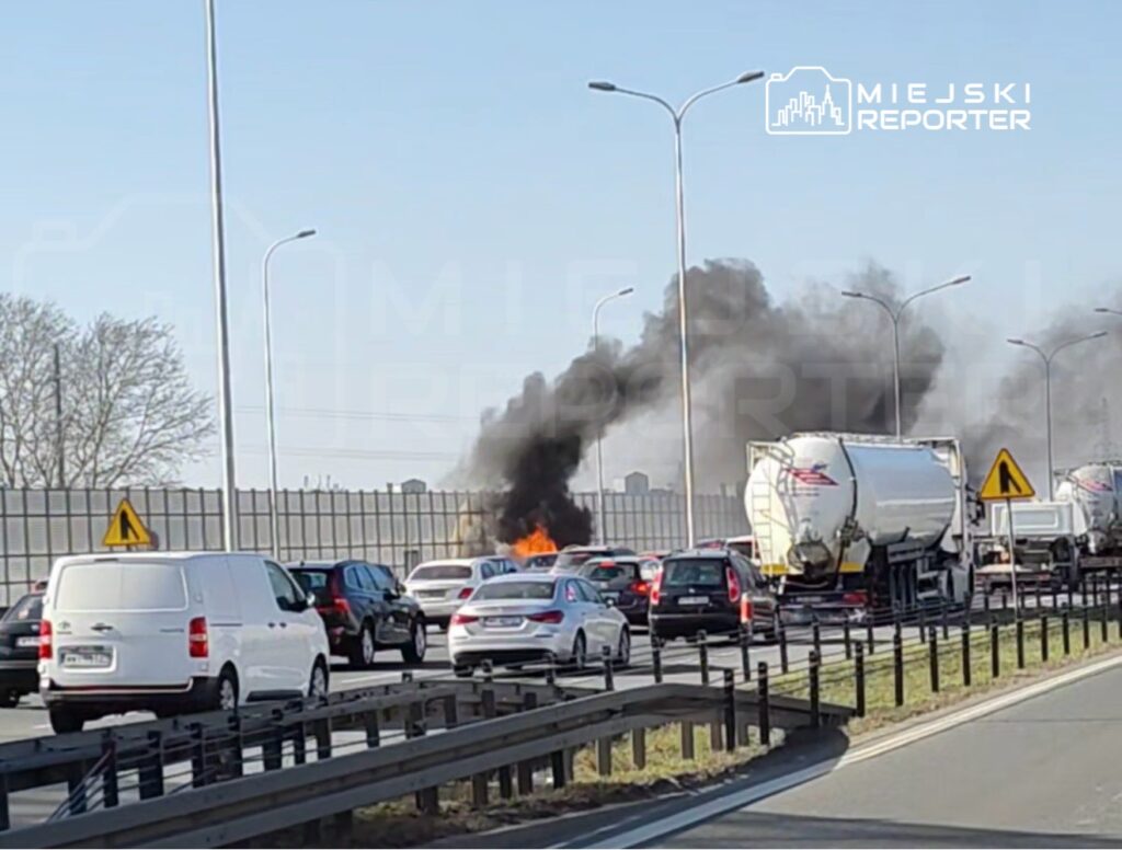 Samochody stoją w korku na drodze, a w tle widoczny jest pożar wydobywający się z jednego z pojazdów.
