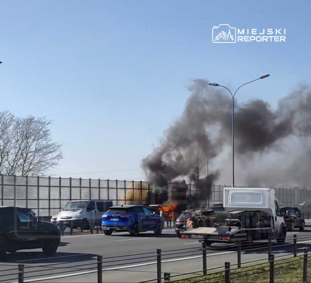 Samochód płonący na autostradzie, wokół widoczne inne pojazdy oraz unoszący się czarny dym.