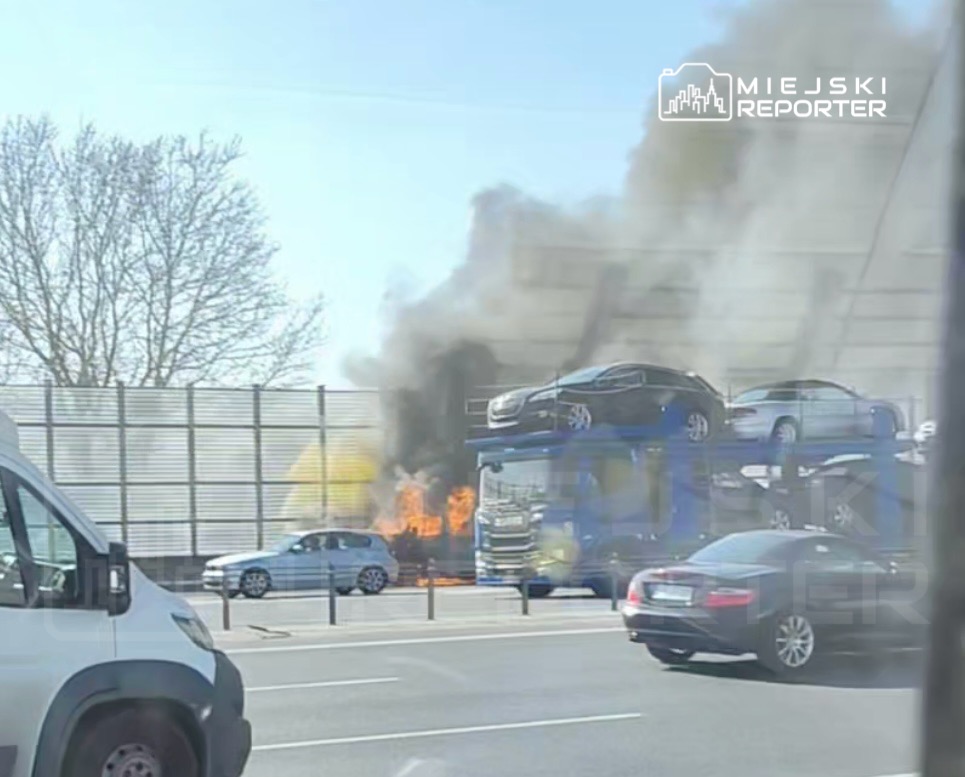 Pożar na ciężarówce transportującej samochody obok ruchliwej drogi, z dymem i płomieniami wydobywającymi się z tyłu.