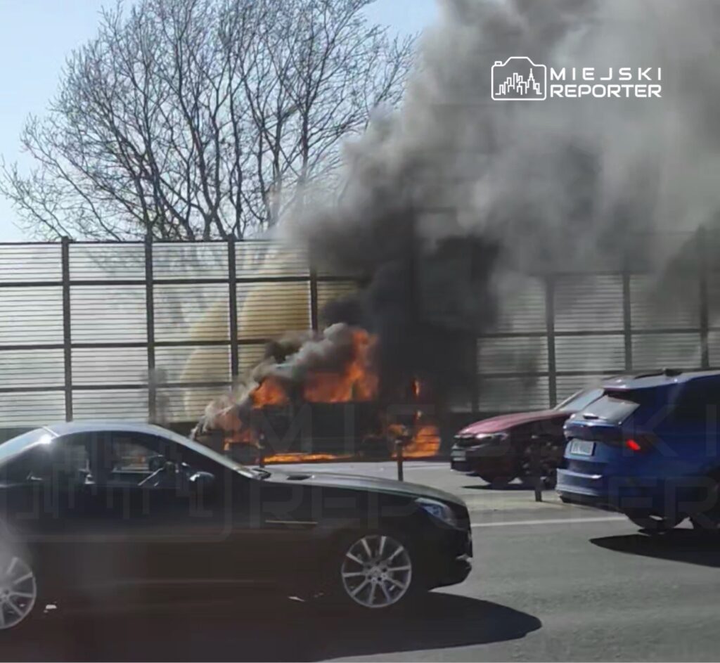 Samochód płonący na poboczu drogi, wokół niego unoszący się czarny dym, a w tle przejeżdżające pojazdy.