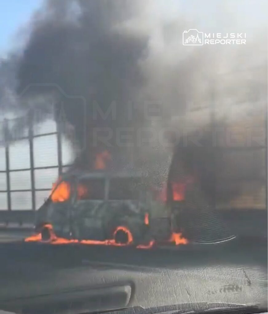 Pojazd w ogniu na drodze, otoczony gęstym dymem, z płomieniami wydobywającymi się z wnętrza.