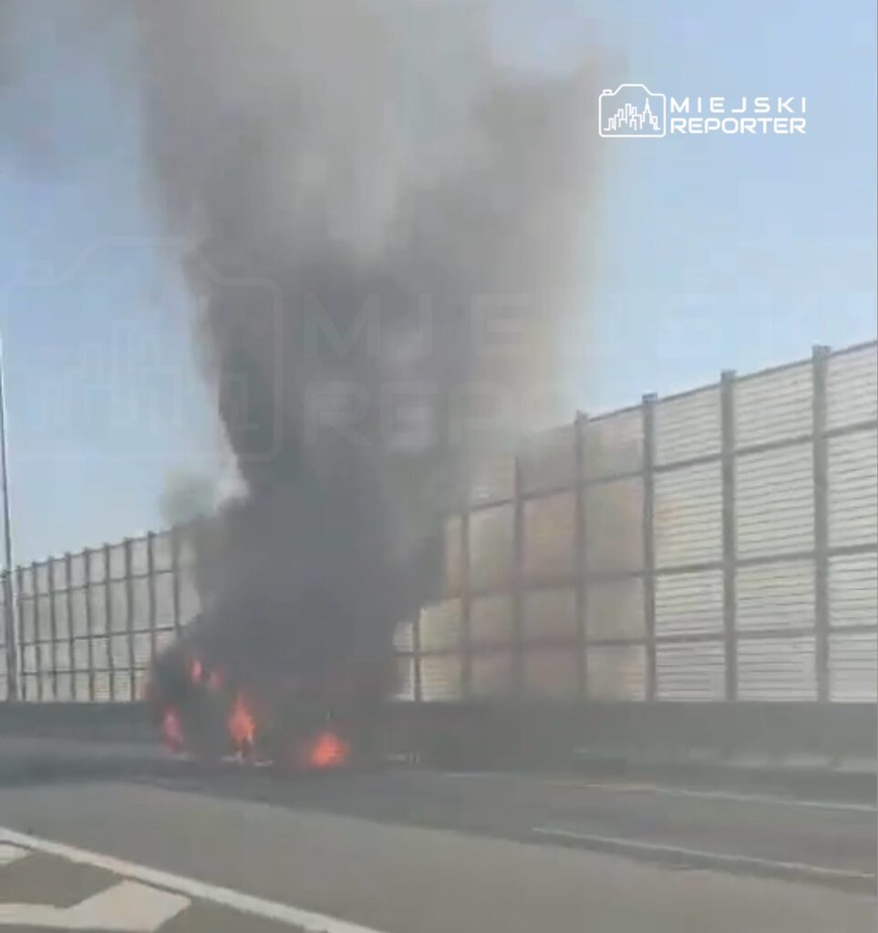 Samochód płonący na pasie drogi, wydobywający czarny dym w pobliżu ogrodzenia akustycznego.