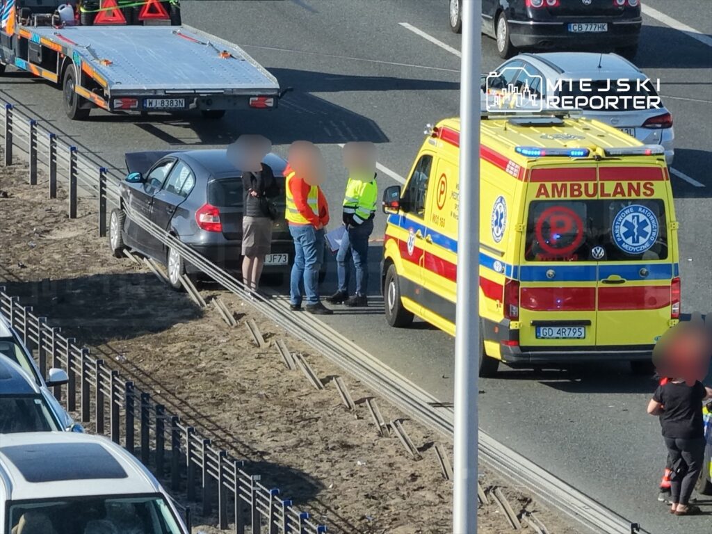 Na poboczu drogi stoi srebrny samochód, obok grupka osób rozmawia z policjantami i ratownikami medycznymi. W tle widoczna karetka.