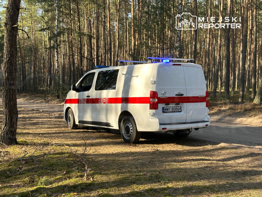 Biały samochód straży z niebieskimi światłami zaparkowany w lesie, obok leśnej drogi.