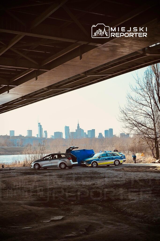 Dwa pojazdy, w tym radiowóz policyjny, stoją na brzegu rzeki, z miejskim krajobrazem w tle.