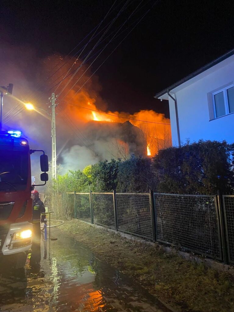Strażak w hełmie obsługuje wąż gaśniczy obok palącego się budynku w nocy, otoczonego przez gęste zarośla.