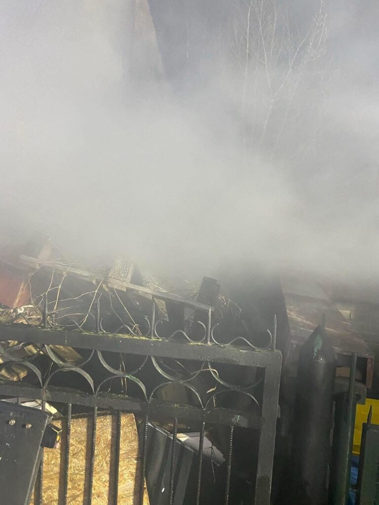 Gęsty dym unoszący się nad zniszczonym ogrodzeniem, w tle widoczne fragmenty zniszczonej konstrukcji.