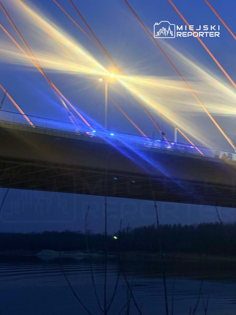 Most z oświetleniem w nocy, z niebieskimi światłami radiowozów na jezdni, w tle woda i zarysy drzew.