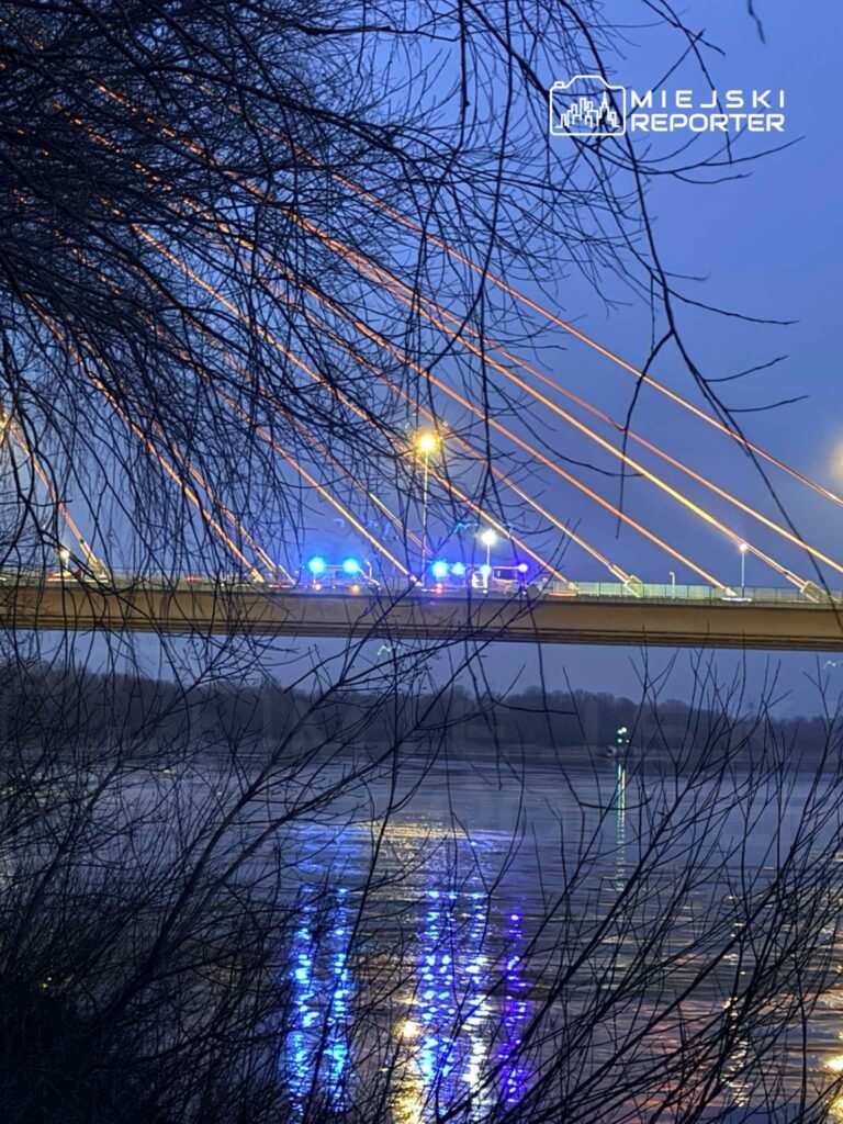 Niebieskie światła radiowozów odbijają się w wodzie pod mostem, otoczonym gałęziami drzew w wieczornym świetle.