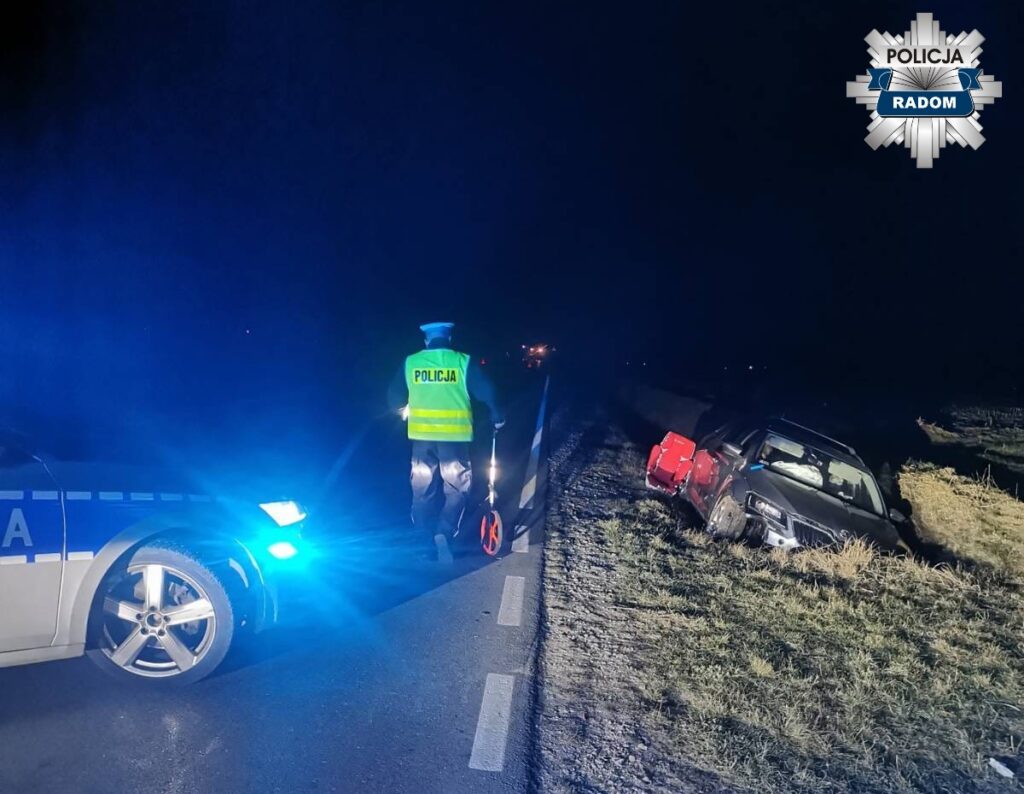 Policjant w kamizelce odblaskowej mierzy odległość przy uszkodzonym samochodzie na poboczu drogi w nocy.