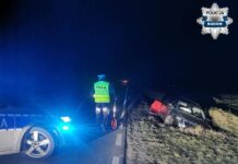 Policjant w kamizelce odblaskowej mierzy odległość przy uszkodzonym samochodzie na poboczu drogi w nocy.