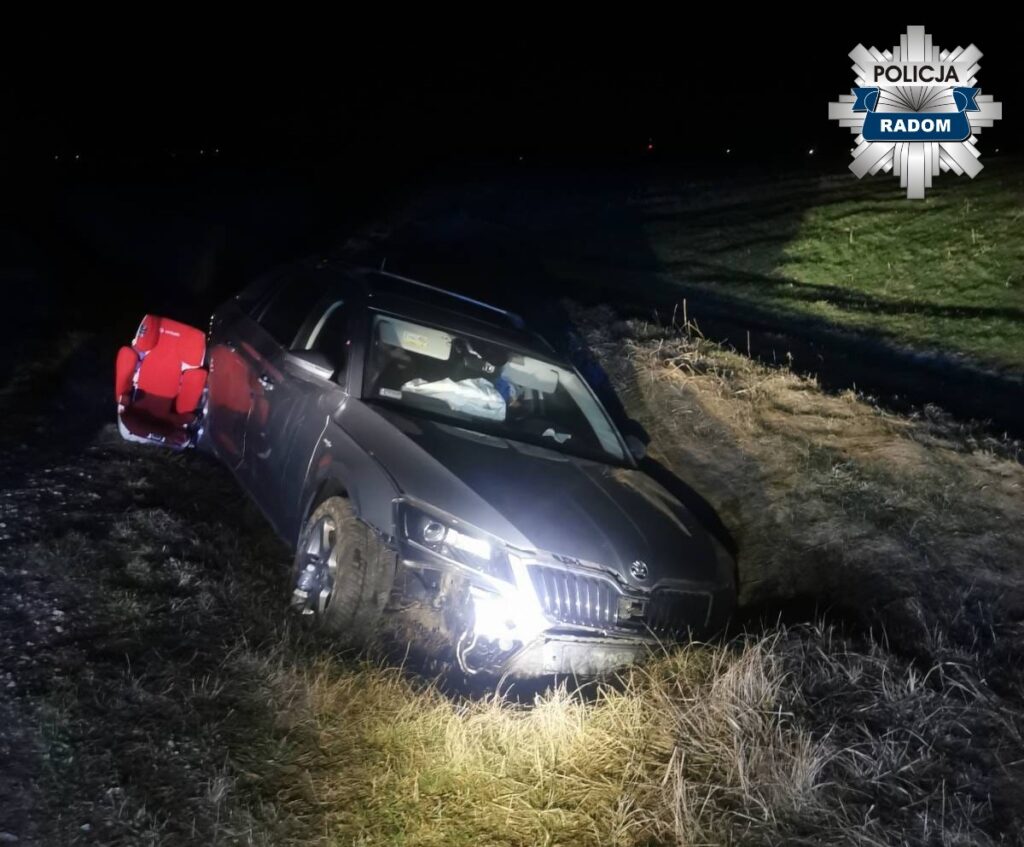 Czarny samochód osobowy stoi na poboczu drogi, z otwartymi drzwiami i fotelikiem dziecięcym na zewnątrz, w nocy.