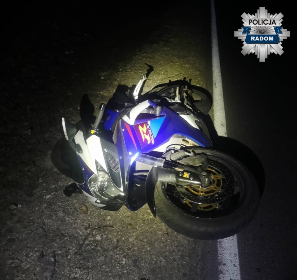 Motocykl leżący na boku przy drodze, oświetlony latarką, w ciemnym otoczeniu.