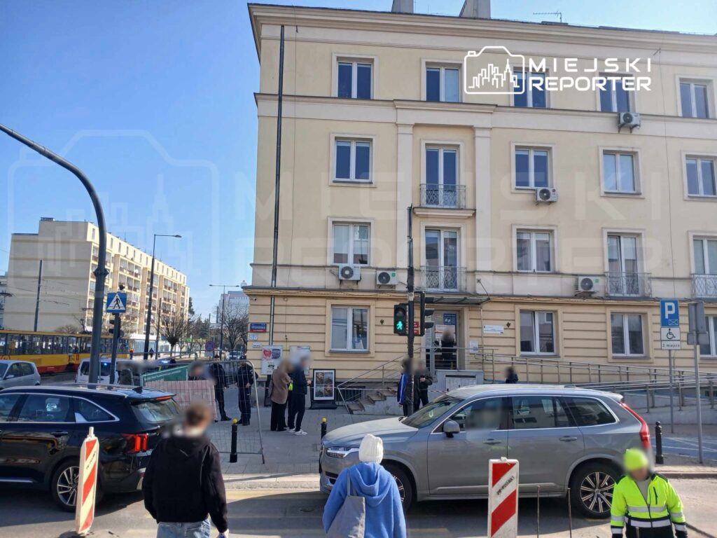 Grupa osób stoi na chodniku przed nowoczesnym budynkiem mieszkalnym, obok zielonego światła dla pieszych.