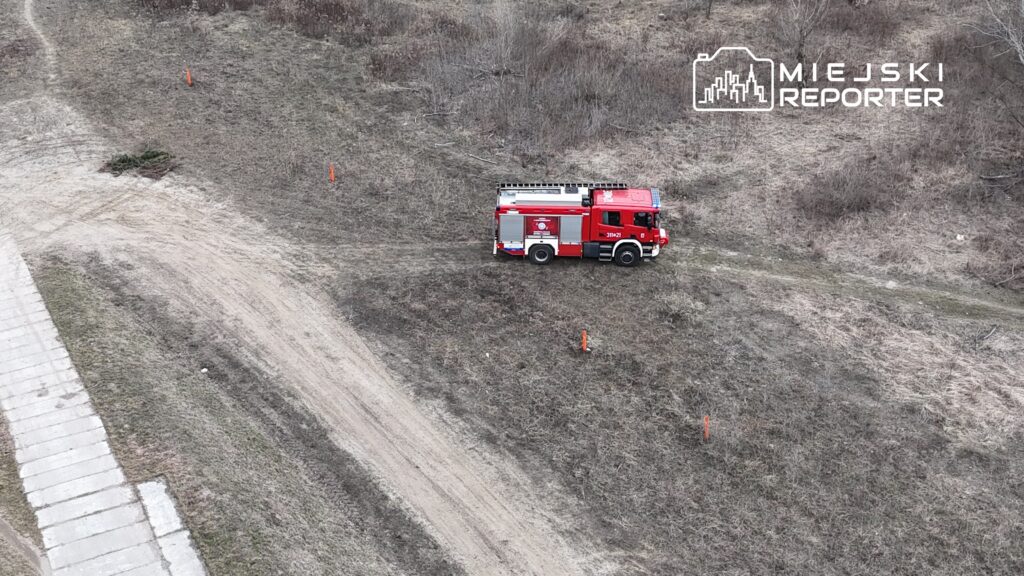 Czerwony wóz strażacki stoi na wyboistej drodze w otoczeniu trawy i zarośli, obok pomarańczowe słupki.