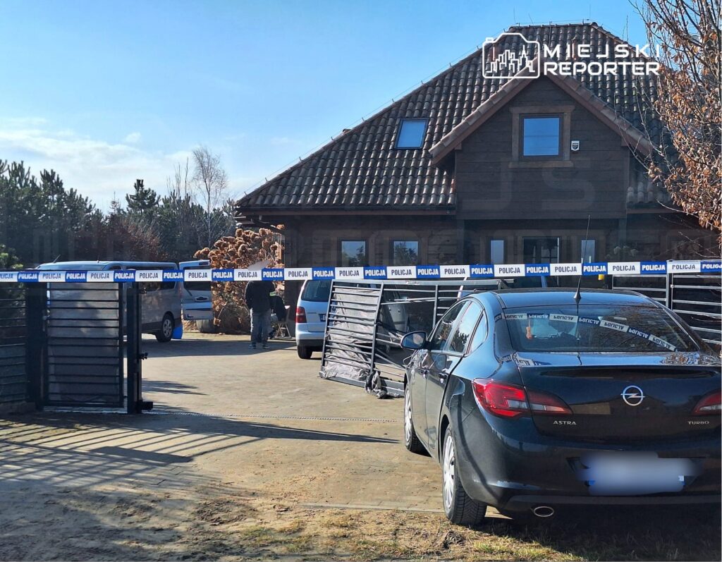 Mężczyzna w pobliżu bramy z taśmą policyjną, obok zaparkowane samochody w otoczeniu domu jednorodzinnego.