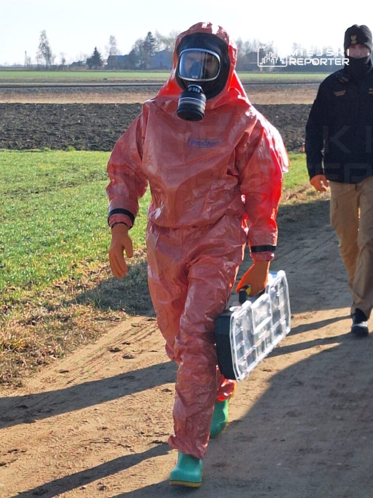 Mężczyzna w kombinezonie ochronnym i masce gazowej idzie po polu, trzymając przezroczystą walizkę.