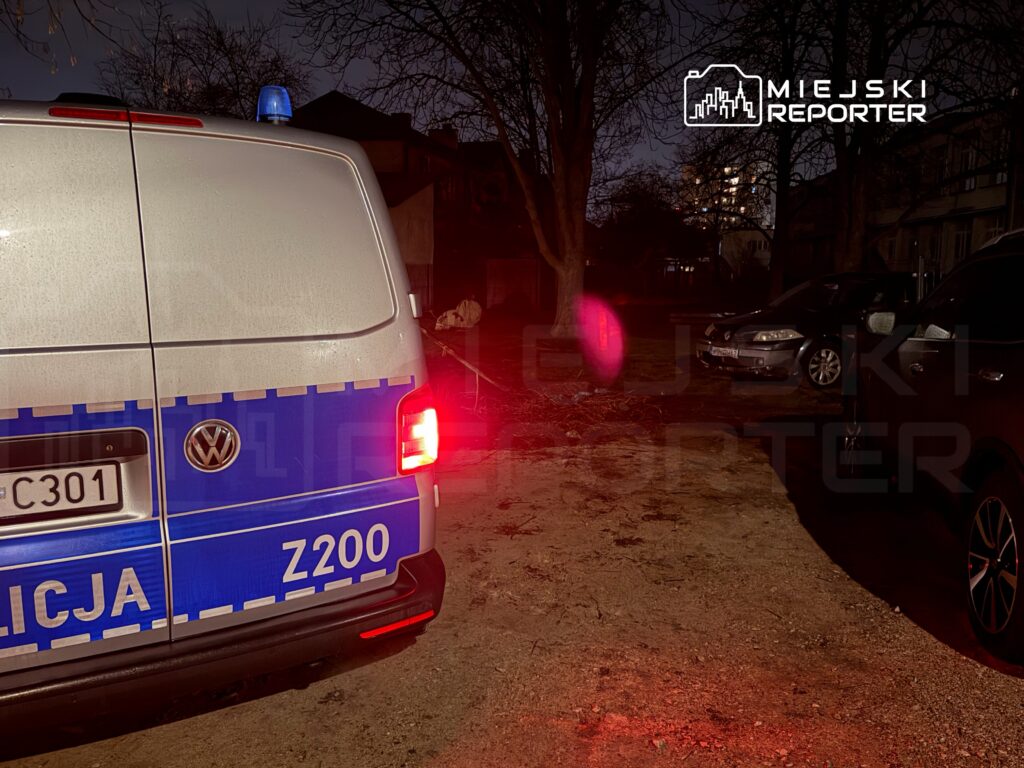 Policyjny samochód z niebieską sygnalizacją zaparkowany w ciemnej uliczce, w otoczeniu zaparkowanych pojazdów.
