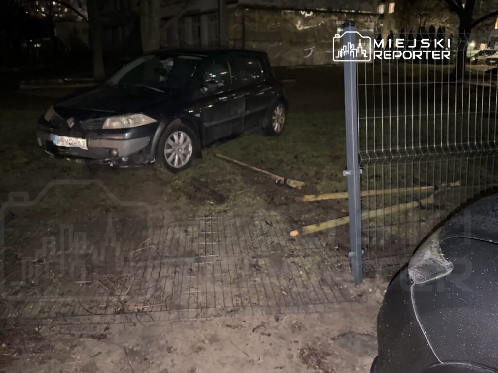 Czarny samochód osobowy zaparkowany na trawie obok ogrodzenia, w tle widoczna ciemna przestrzeń nocna.