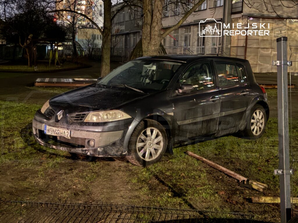Czarny samochód osobowy zaparkowany na trawniku obok ścieżki w miejskim otoczeniu, w tle widoczne budynki.