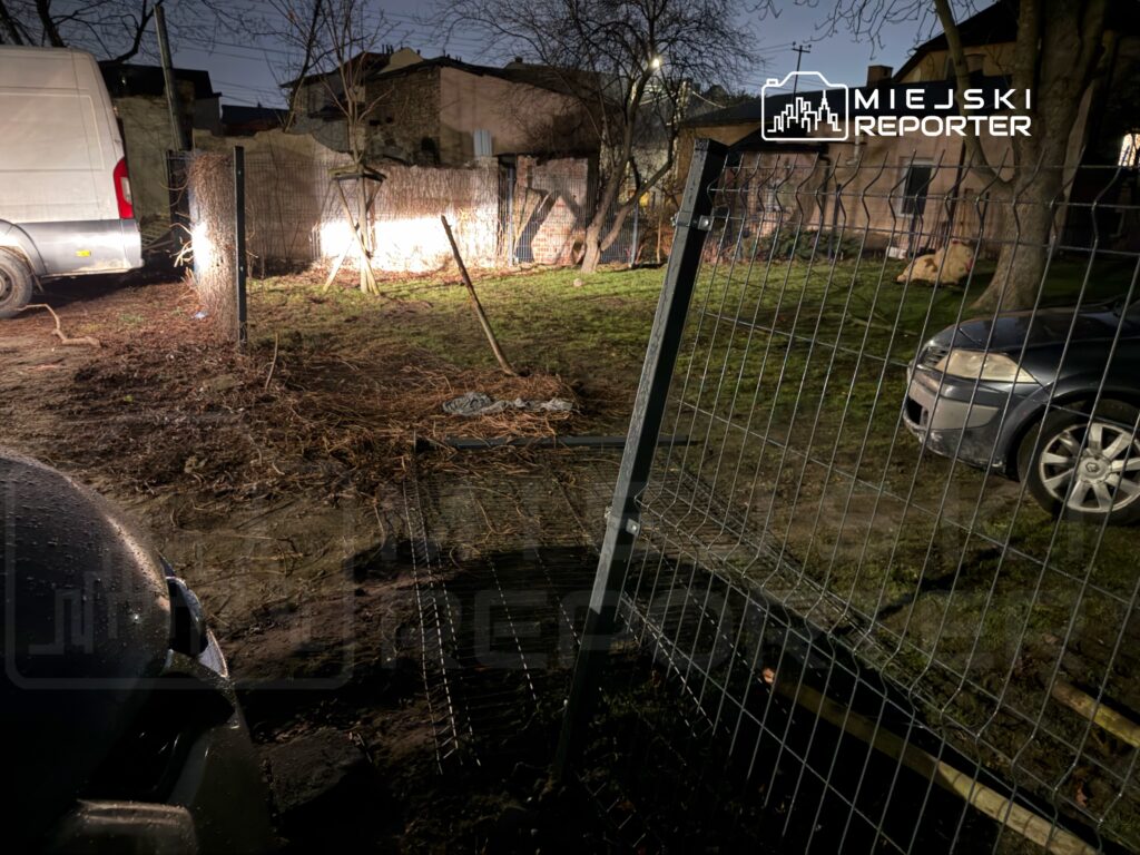 W nocy na podwórku, oświetlonym przez reflektory, widać van oraz dwa zaparkowane samochody obok uszkodzonego ogrodzenia.
