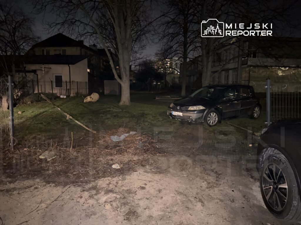 Samochód zaparkowany na trawie obok budynku mieszkalnego w nocy, otoczony drzewami i zaroślami.
