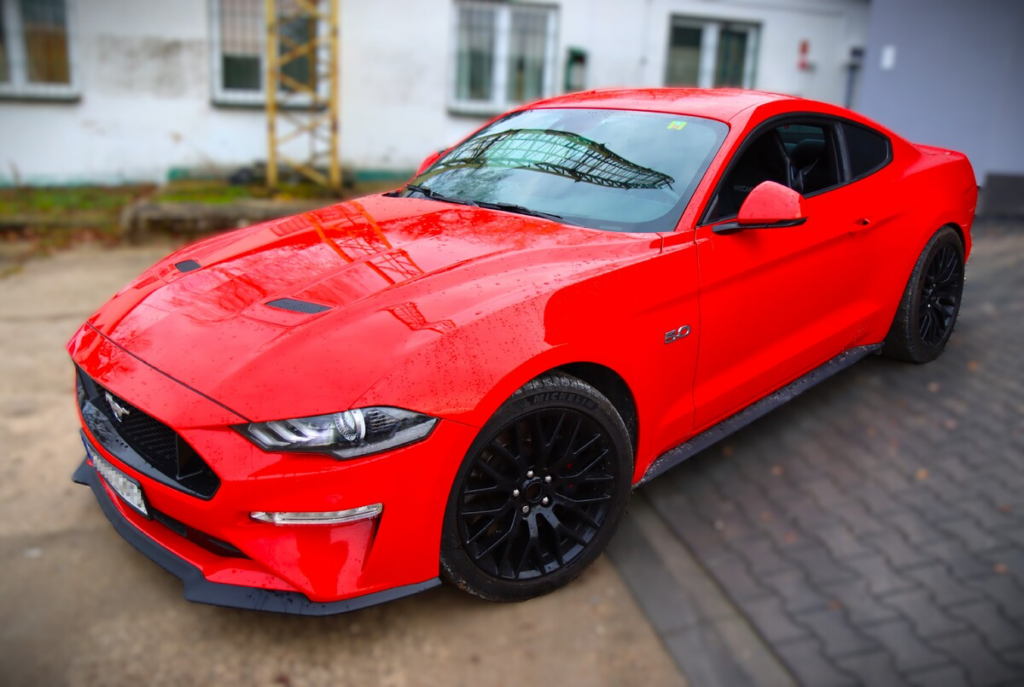 Czerwony sportowy samochód Mustang zaparkowany na brukowanej nawierzchni w otoczeniu budynków.