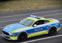 Policyjny samochód sportowy z niebiesko-żółtym malowaniem, zatrzymany na torze wyścigowym, z kierowcą w okularach przeciwsłonecznych.