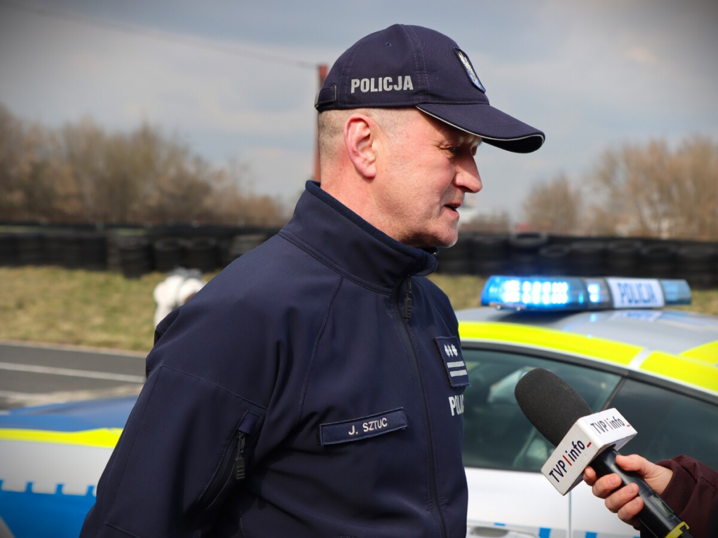 Policjant w mundurze rozmawia z dziennikarzem przy radiowozie z włączonymi sygnałami świetlnymi.