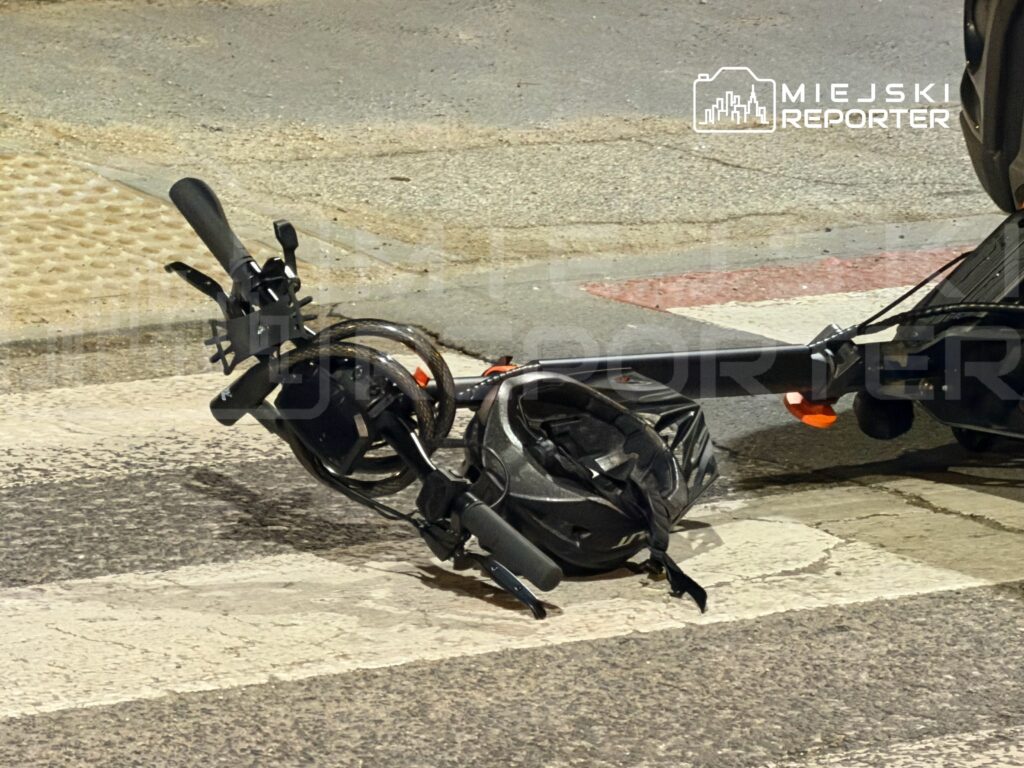 Złamana hulajnoga leży na pasie drogowym obok torów tramwajowych, w nocy przy oświetlonej ulicy.