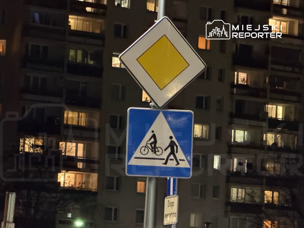 Znaki drogowe informujące o strefie dla pieszych i rowerzystów, w tle oświetlone budynki mieszkalne.