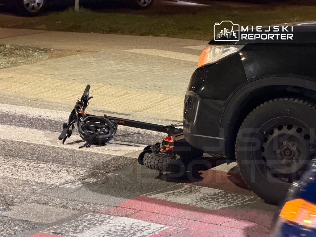 Złamana hulajnoga leży na pasach, obok czarnego SUV-a, w nocy na miejskiej ulicy.