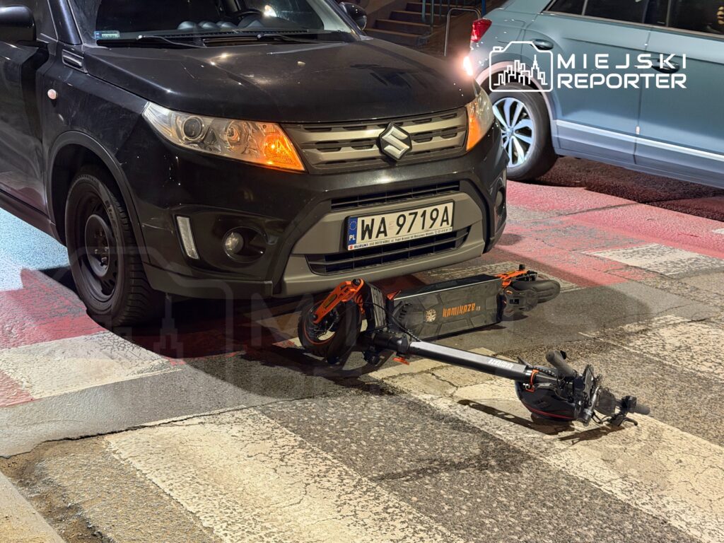 Czarna osobówka zaparkowana na przejściu dla pieszych, z elektryczną hulajnogą unieruchomioną pod przednim zderzakiem.