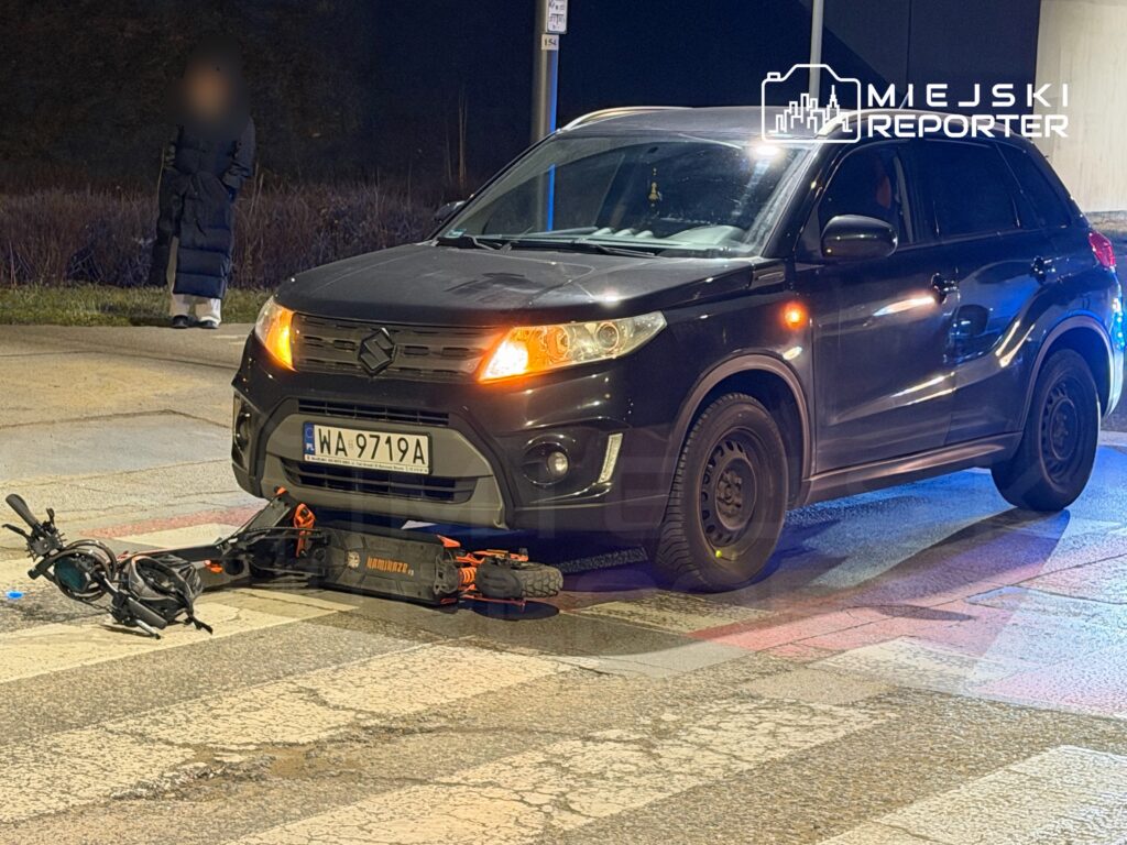 Czarny samochód osobowy stoi na przejściu dla pieszych, a pod jego przednim kołem leży uszkodzona hulajnoga elektryczna.