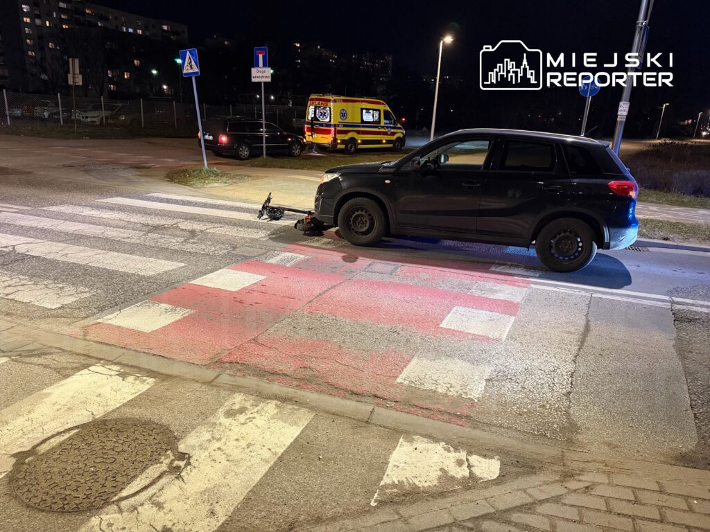 Czarny samochód osobowy stoi na przejściu dla pieszych w nocy, obok widać ambulans i znaki drogowe.
