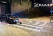 Czarny SUV zatrzymany na przejściu dla pieszych, obok leżący skuter elektryczny, w tle oświetlony chodnik.