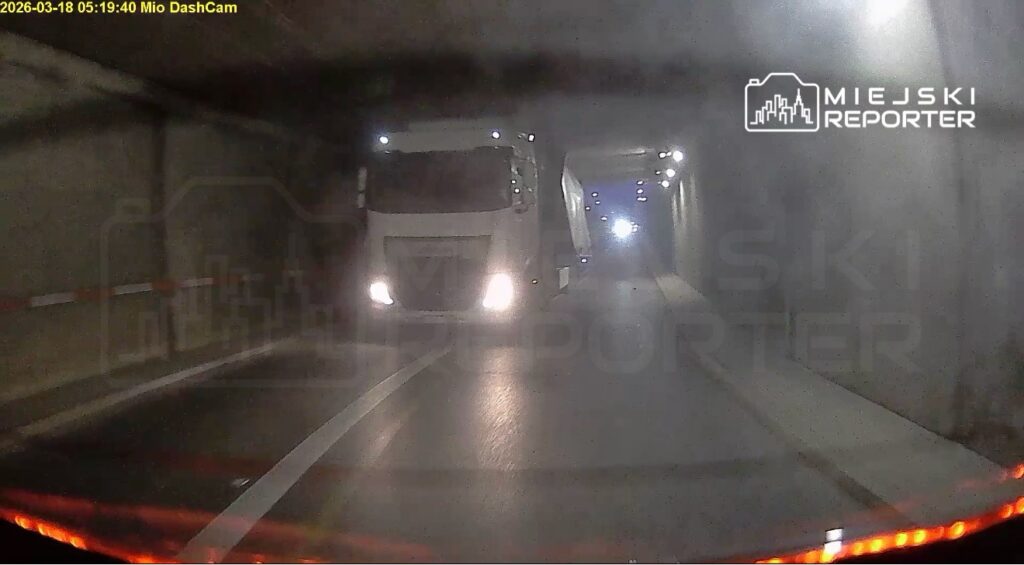 Ciężarówka z naczepą przejeżdża przez oświetlony tunel, mijając inne pojazdy w ciemnym otoczeniu.